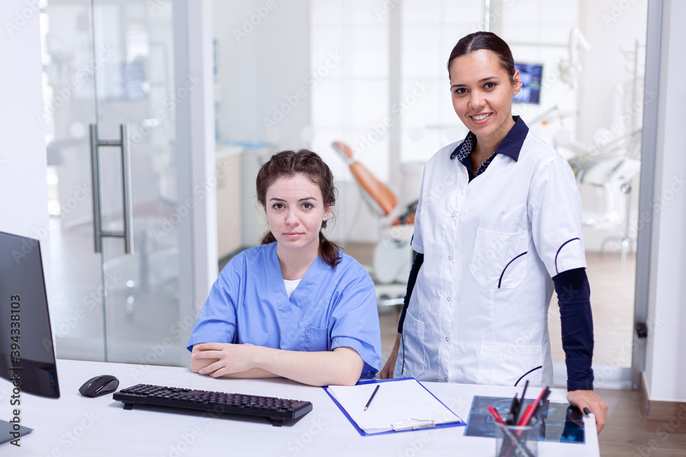 Obraz premium Medical team in dental office smiling at camera wearing uniform. Stomatology assistant and teeth doctor discussing in reception of dental office