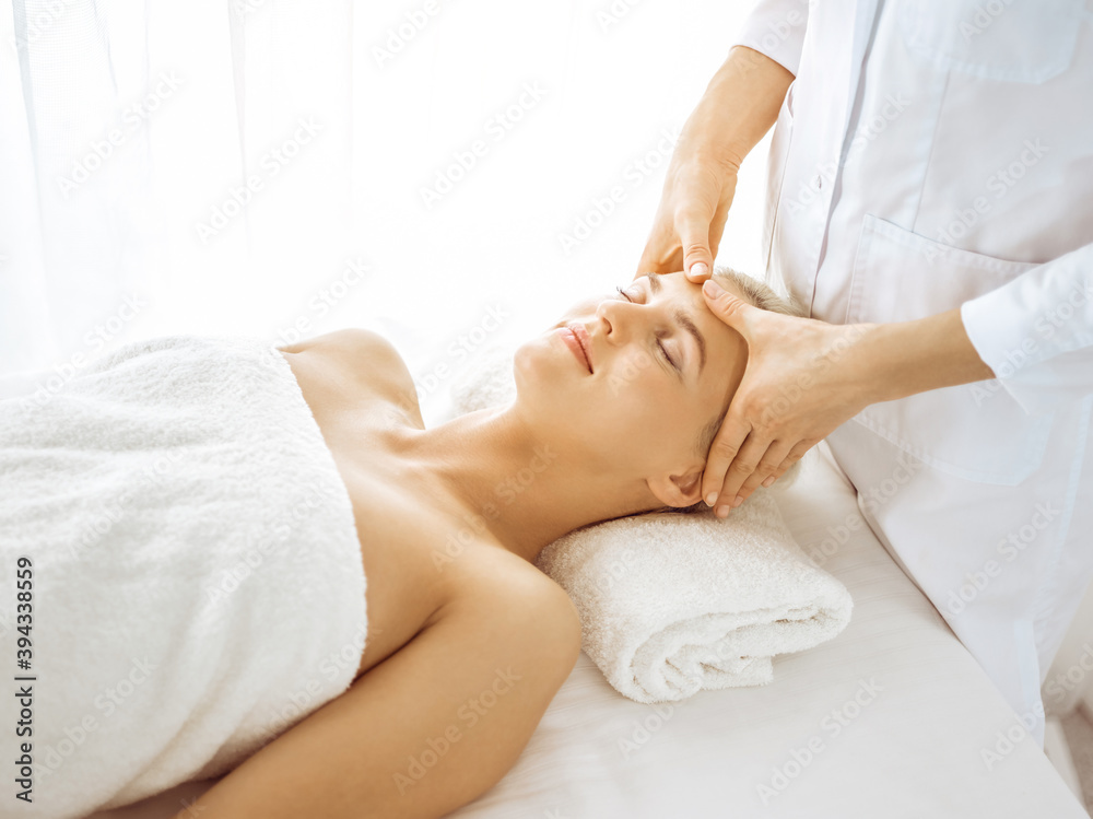 Beautiful blonde woman enjoying facial massage with closed eyes in sunny spa center. Relaxing treatment in medicine and beauty concepts Beautiful blonde woman enjoying facial massage with closed eyes in sunny spa center. Relaxing treatment in medicine and beauty concepts