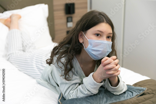 A teenage girl in bed lying down watching an interesting movie in a mask and very thoughtful. Quarantine