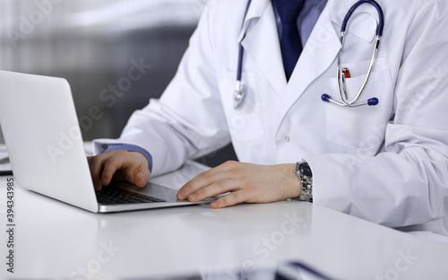 Unknown male doctor sitting and working with laptop in clinic at his working place, close-up. Young physician at work. Perfect medical service, medicine concept