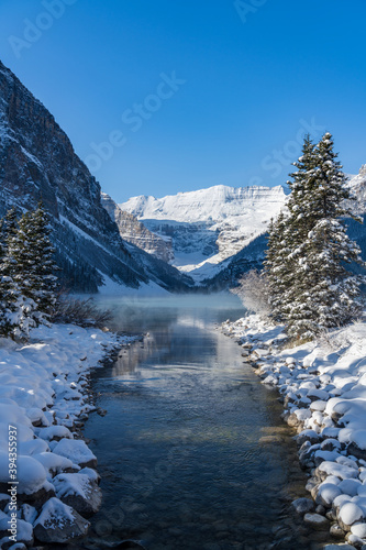 Lake Louise in early winter sunny day morning. Mist floating on turquoise color water surface. Clear blue sky, snow capped mountains in background. Beautiful natural landscape in Banff National Park. © Shawn.ccf