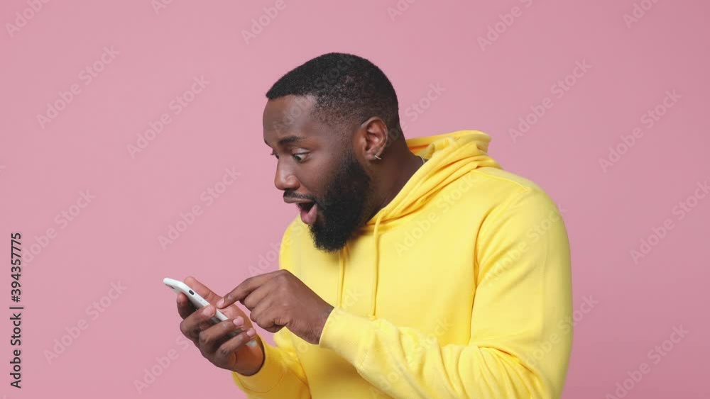 Excited shocked young african american man 20s wearing yellow hoodie posing isolated on pastel pink color background studio. People lifestyle concept. Using mobile cell phone typing browsing say wow Excited shocked young african american man 20s wearing yellow hoodie posing isolated on pastel pink color background studio. People lifestyle concept. Using mobile cell phone typing browsing say wow