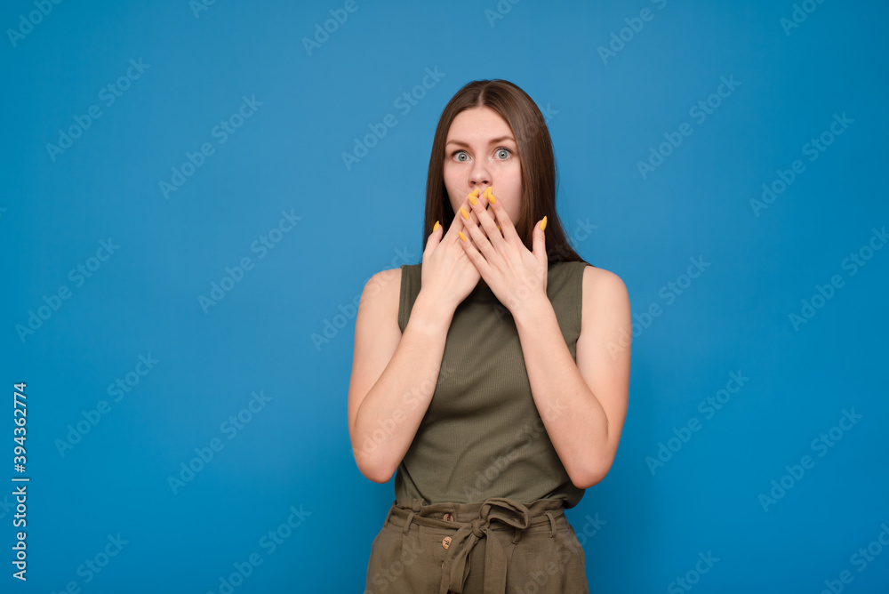 Young beautiful caucasian woman in a green t-shirt on an isolated blue background surprised. Covers his face with his hands Young beautiful caucasian woman in a green t-shirt on an isolated blue background surprised. Covers his face with his hands