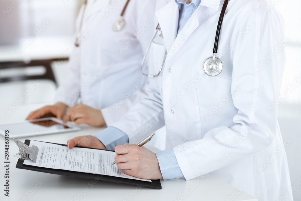 Unknown woman-doctors at work with patient at the background. Female physicians filling up medical documents or prescription while standing in sunny hospital reception desk, close-up. Health care Unknown woman-doctors at work with patient at the background. Female physicians filling up medical documents or prescription while standing in sunny hospital reception desk, close-up. Health care