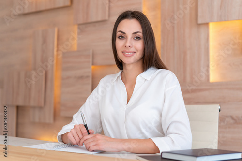 Office manager woman in white shirt, beautiful smile, sitting to table with notebook in wooden office interior, side view. Friendly office secretary looking at the camera. Concept of work