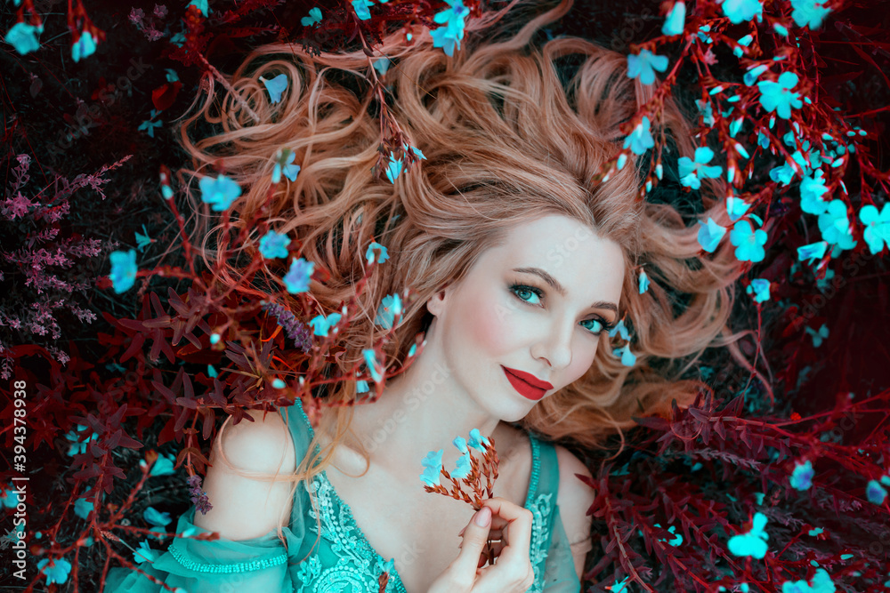beautiful happy fantasy elf woman with blond hair lies dreams on grass,  blooming meadow, blue flowers. Portrait smiling face, red lips elegant  makeup. Romantic Girl princess resting enjoying life Stock Photo