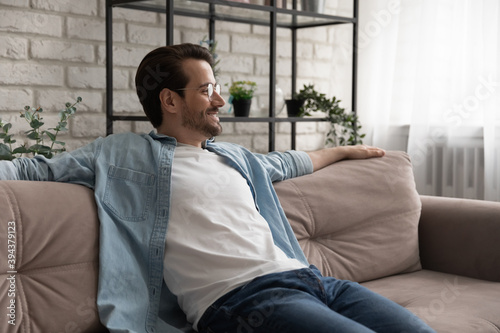 Smiling young handsome man in glasses relaxing on comfortable couch, breathing fresh air indoors, enjoying stress free leisure weekend time alone in living room, watching tv series, domestic pastime
