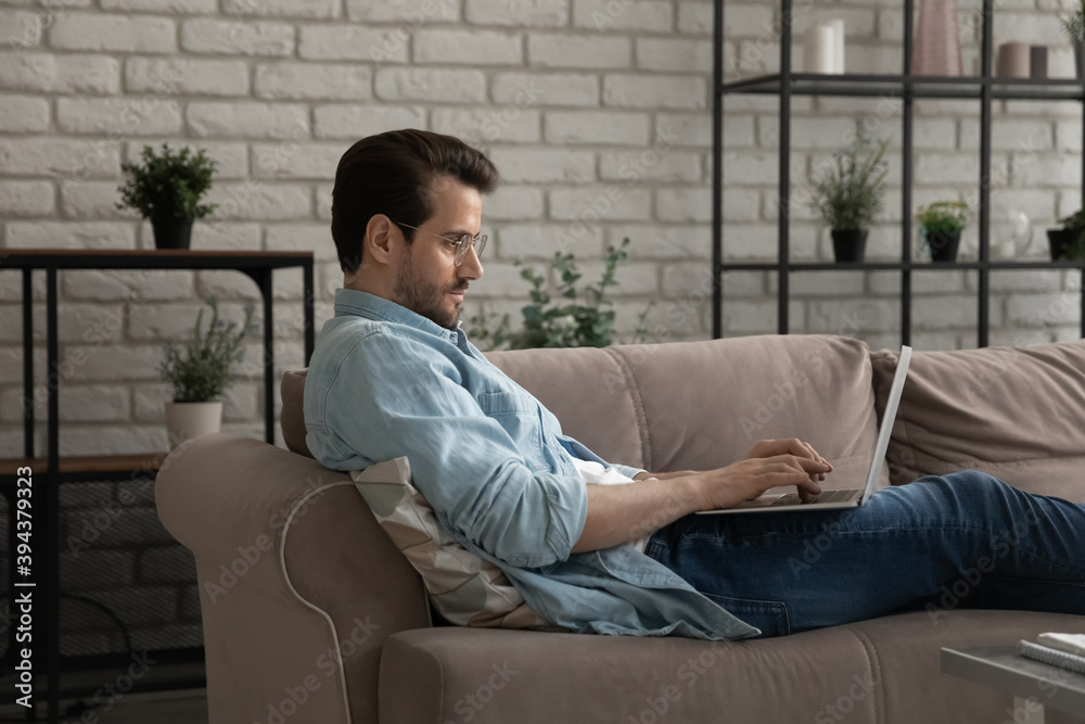 Relaxed young man lying on sofa with computer on laps, involved in web surfing information, communicating distantly with friends in social network, spending leisure free time online, tech addiction.
