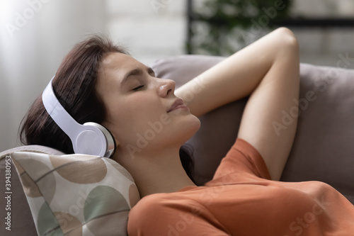 Head shot peaceful young woman lying on pillows on comfortable sofa, listening music or positive affirmations in modern headphones with closed eyes, relaxing alone at home, domestic hobby concept. photo