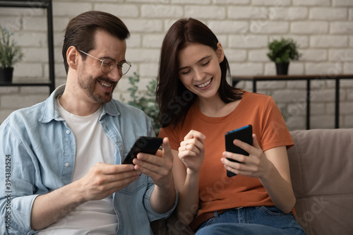 Addicted to technology happy caucasian young married family couple sitting on comfortable couch, laughing at funny video photo content in social networks, enjoying using smartphones together at home.