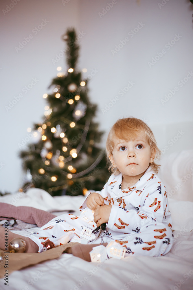 the child's first Christmas. a pensive little boy is lying in his holiday pyjamas on a bed against the background of a brightly decorated fir tree with a Golden light garland. the child's first Christmas. a pensive little boy is lying in his holiday pyjamas on a bed against the background of a brightly decorated fir tree with a Golden light garland.