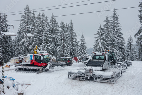 Snow ploughs with caterpillar tracks are parked up in a european ski resort having been out clearing snow in a ski resort.Ski lift chairs and pine forest in the winter scene.