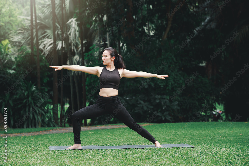 Asian woman practicing yoga in Warrior Pose (Virabhadrasana) on the mat in outdoor park. Asian woman practicing yoga in Warrior Pose (Virabhadrasana) on the mat in outdoor park.