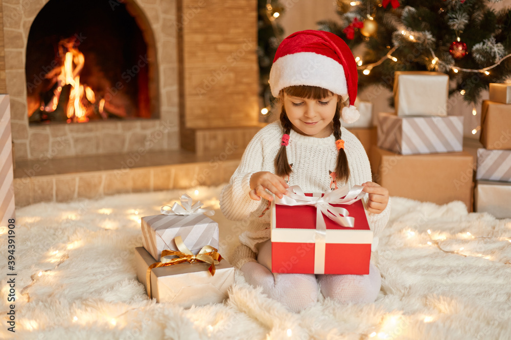 Little girl opening christmas present, keeping fingers on ribbons, looking at gift box with smile, child wearing santa claus hat posing on floor near fireplace and xmas tree.