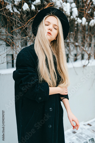 Stylish beautiful blonde girl in black clothes and hat posing near fence in the street