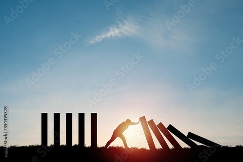 Silhouette man pushing rectangle  block which falling to stop dominos others rectangle standing with blue sky. Risk and crisis management concept.