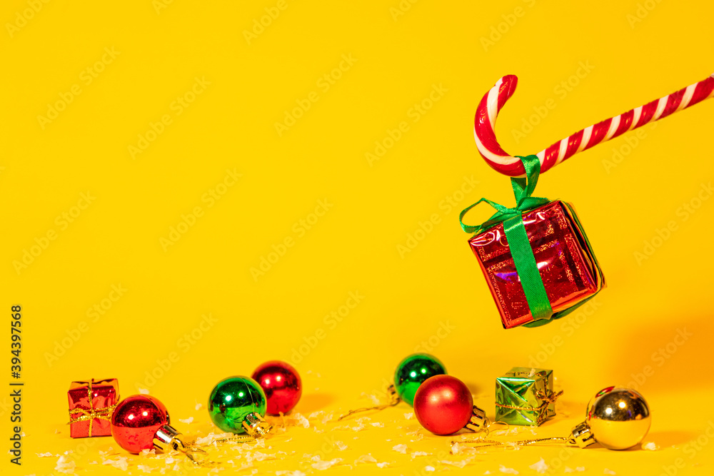 Fototapeta premium Candy cane lollipop holding a gift box with a Christmas present on a yellow background, Christmas sweets with new year decorations