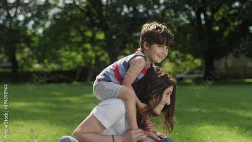 A young son sitting on the shoulders of his young mom while sitting on the grass in a park