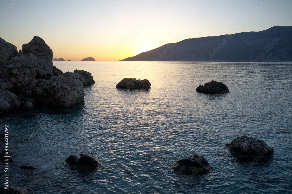 View of sunset at the seaside. Calm ocean with orange sun on the horizon. Large stone boulders on the sea shore.