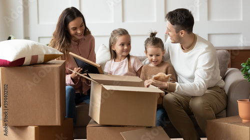 Family with 6s kids moved to new house sitting on couch in living room unpacking personal belongings from cardboard boxes chatting enjoy relocation day. Bank loan, happy homeowners unbox stuff concept