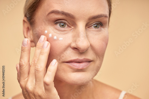 Close up of beautiful mature woman applying anti-aging cream under the eyes, isolated on beige background