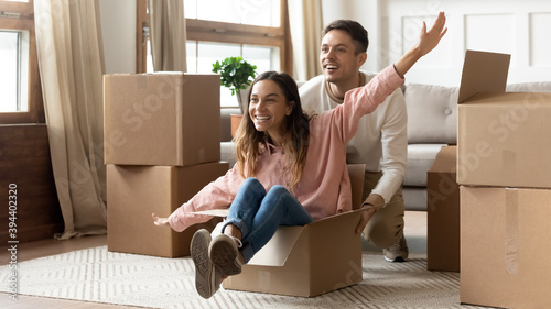 Overjoyed husband pushing playful wife seated inside of cardboard box raised overstretched arms, family having fun celebrate relocation to new house. Moving day celebration, bank loan, lease concept