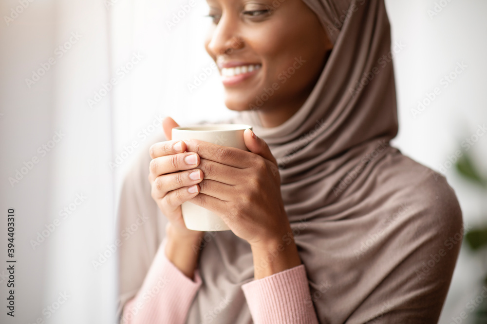 Cropped view of happy black woman in hijab warming her hands on cup of hot coffee near window at home Cropped view of happy black woman in hijab warming her hands on cup of hot coffee near window at home