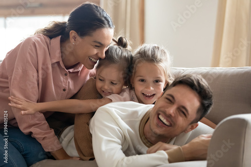 Portrait of happy carefree loving full family with little daughters relaxing at home, fun and love concept. Two cute girls piggyback father, people spend weekend together lying on couch in living room