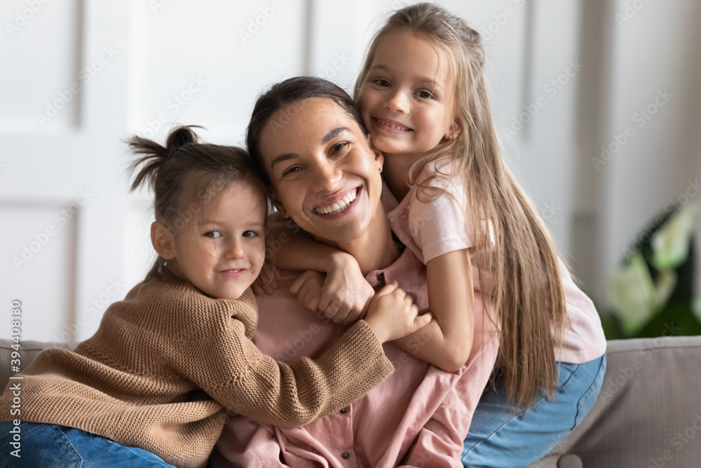Little 6s 7s beautiful daughters hug young loving mommy, family sitting on couch in living room embracing smiling looking at camera posing for family portrait. Happy Mother Day celebration concept Little 6s 7s beautiful daughters hug young loving mommy, family sitting on couch in living room embracing smiling looking at camera posing for family portrait. Happy Mother Day celebration concept