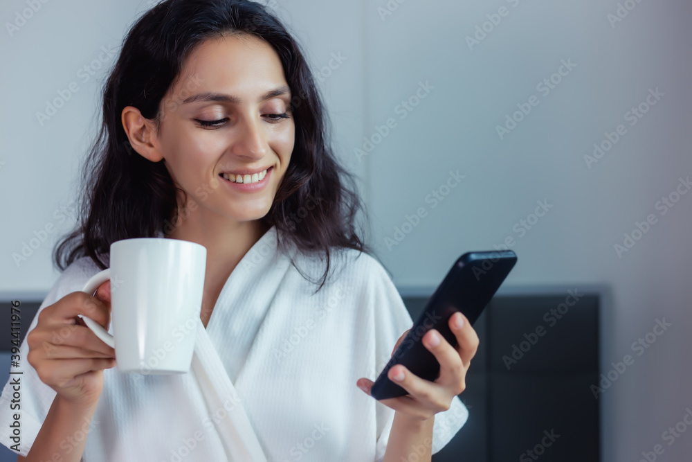 Beautiful smiling woman using smart phone and enjoying hot coffee in the morning at bedroom. Young woman in bed with smart phone and cup of coffee in hand. Happy female using mobile phone and internet Beautiful smiling woman using smart phone and enjoying hot coffee in the morning at bedroom. Young woman in bed with smart phone and cup of coffee in hand. Happy female using mobile phone and internet