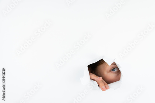 A woman peeks through a hole in a white paper wall with one eye.