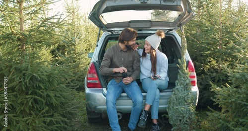Good-looking amorous happy smiling couple resting in car's trunk after buying fir tree in forestry and enjoying joint conversation,front view