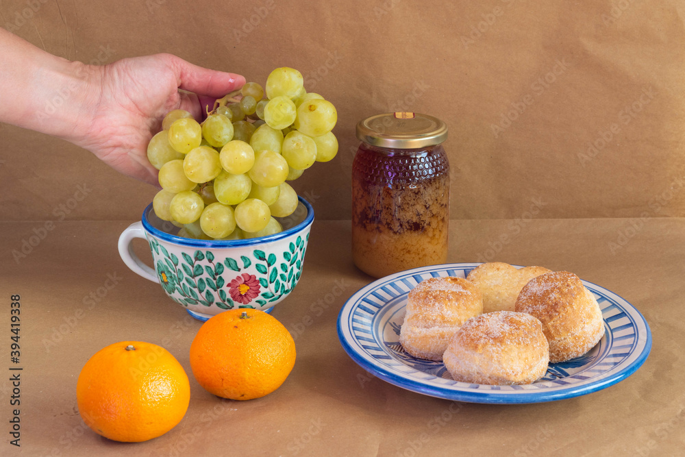 Woman's hand catching a grape cluster surrounded by some traditional Spanish cakes, a pot of honey, tangerines presented in traditional hand-painted ceramic. Woman's hand catching a grape cluster surrounded by some traditional Spanish cakes, a pot of honey, tangerines presented in traditional hand-painted ceramic.