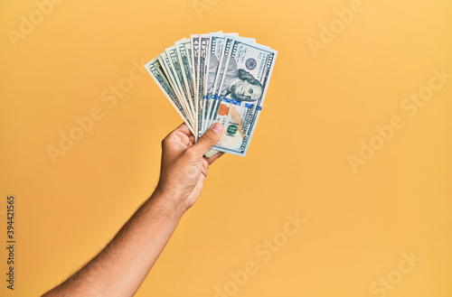Hand of hispanic man holding american dollars banknotes over isolated yellow background.