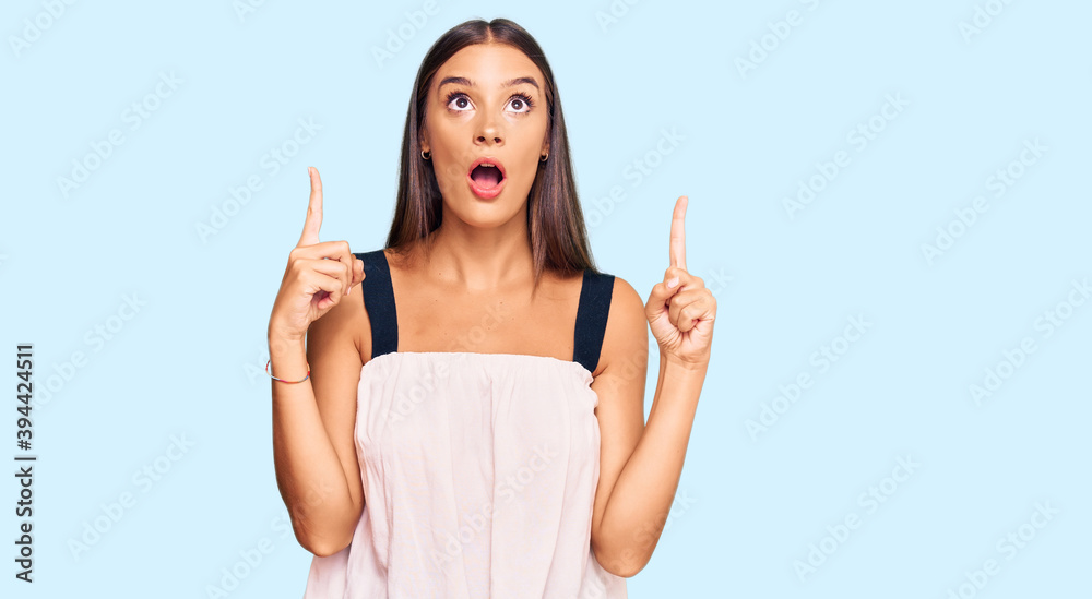 Young hispanic woman wearing casual clothes amazed and surprised looking up and pointing with fingers and raised arms. Young hispanic woman wearing casual clothes amazed and surprised looking up and pointing with fingers and raised arms.
