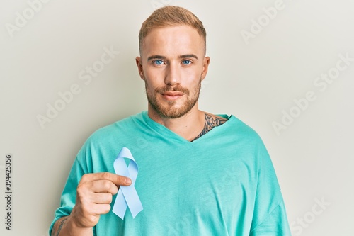 Young caucasian man holding blue ribbon thinking attitude and sober expression looking self confident