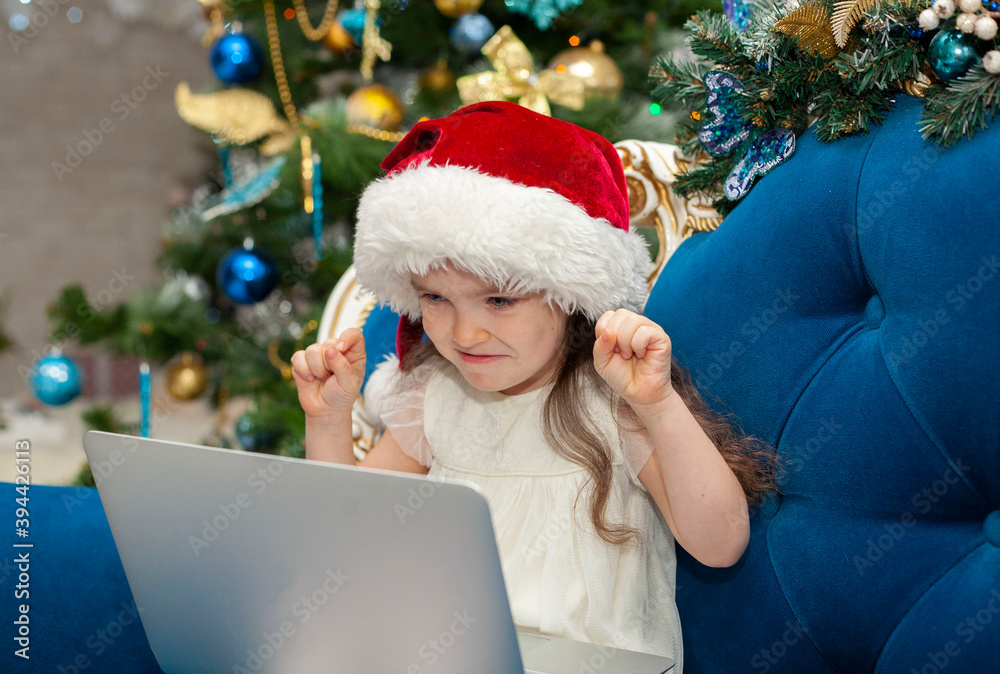 A little girl in a hat of Santa Claus grimaces at the camera. Happy New Year on-line on a laptop. A little girl in a hat of Santa Claus grimaces at the camera. Happy New Year on-line on a laptop.