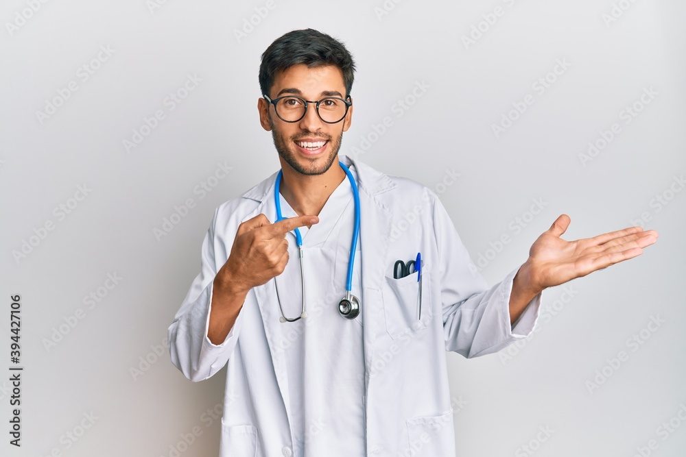 Young handsome man wearing doctor uniform and stethoscope amazed and smiling to the camera while presenting with hand and pointing with finger. Young handsome man wearing doctor uniform and stethoscope amazed and smiling to the camera while presenting with hand and pointing with finger.
