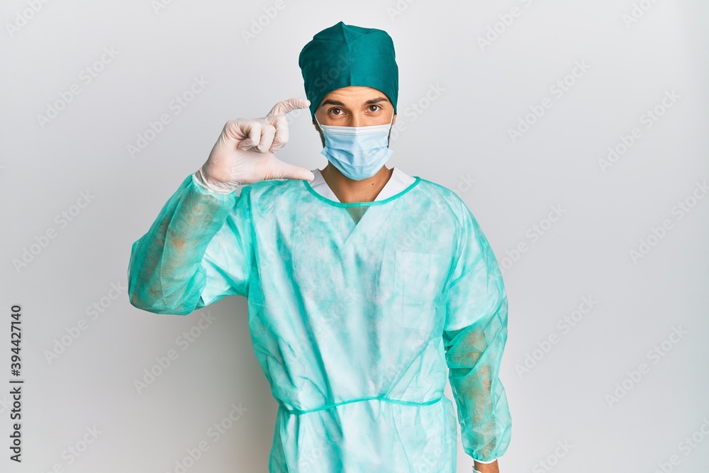 Young handsome man wearing surgeon uniform and medical mask smiling and confident gesturing with hand doing small size sign with fingers looking and the camera. measure concept. Young handsome man wearing surgeon uniform and medical mask smiling and confident gesturing with hand doing small size sign with fingers looking and the camera. measure concept.
