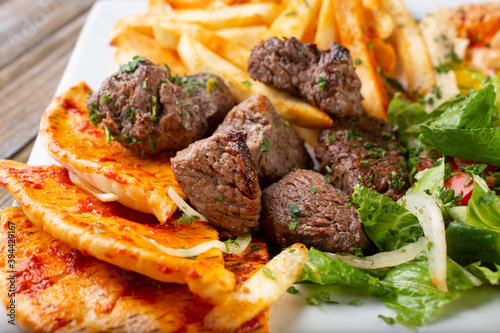 A closeup view of a plate of beef kabob, featuring sides of pita bread and french fries.