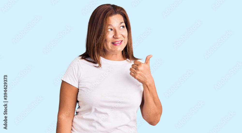Middle age latin woman wearing casual white tshirt smiling with happy face looking and pointing to the side with thumb up. Middle age latin woman wearing casual white tshirt smiling with happy face looking and pointing to the side with thumb up.