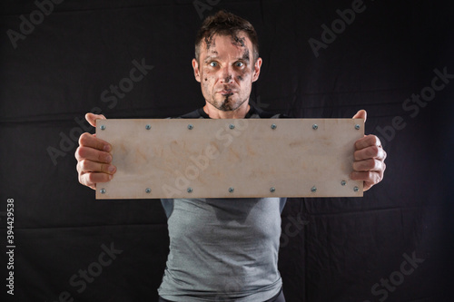 A sporty man holding an empty wooden sign with screws on a black background. Concept for inserting any word