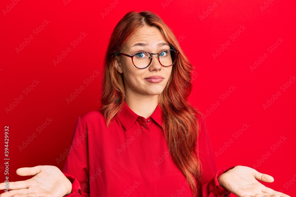 Young beautiful redhead woman wearing casual clothes and glasses over red background clueless and confused expression with arms and hands raised. doubt concept.