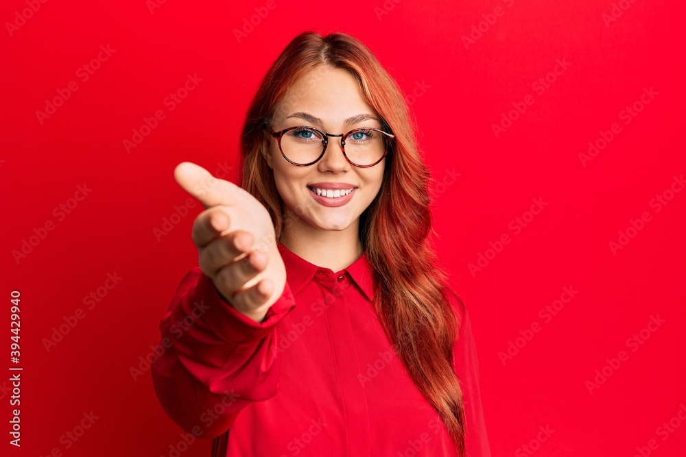 Young beautiful redhead woman wearing casual clothes and glasses over red background smiling friendly offering handshake as greeting and welcoming. successful business.