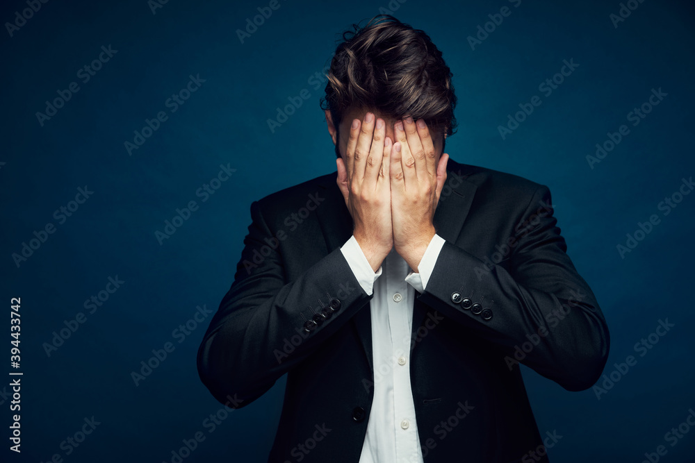 Portrait of depressed businessman hiding head in hands against dark blue background Portrait of depressed businessman hiding head in hands against dark blue background