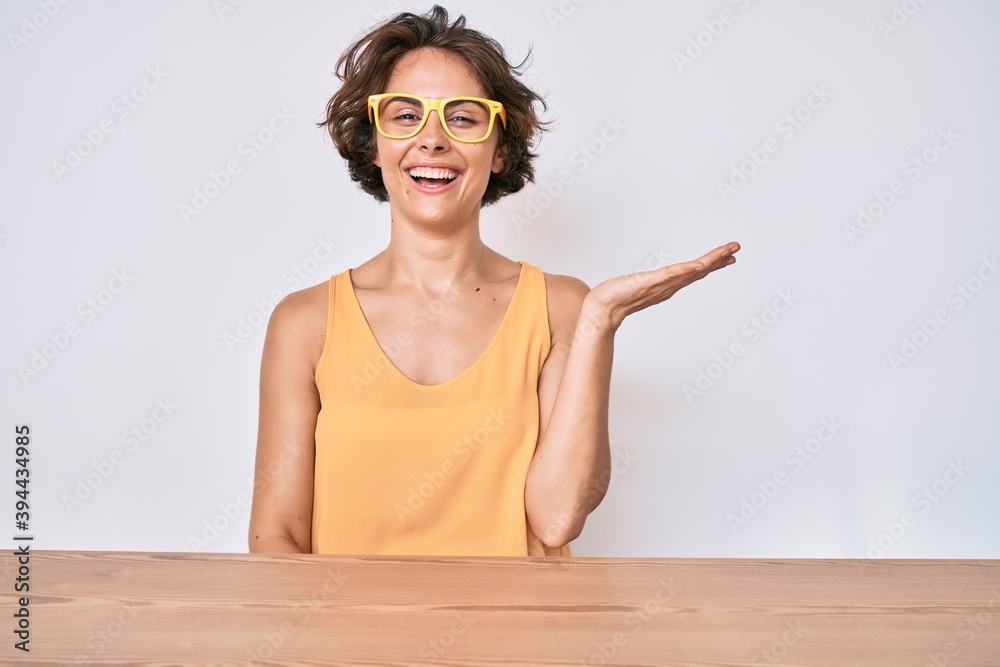 Young hispanic woman wearing casual clothes and glasses sitting on the table smiling cheerful presenting and pointing with palm of hand looking at the camera. Young hispanic woman wearing casual clothes and glasses sitting on the table smiling cheerful presenting and pointing with palm of hand looking at the camera.