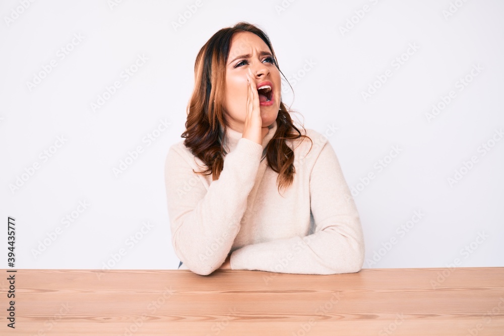 Young beautiful caucasian woman wearing turtleneck sweater sitting on the table shouting and screaming loud to side with hand on mouth. communication concept. Young beautiful caucasian woman wearing turtleneck sweater sitting on the table shouting and screaming loud to side with hand on mouth. communication concept.