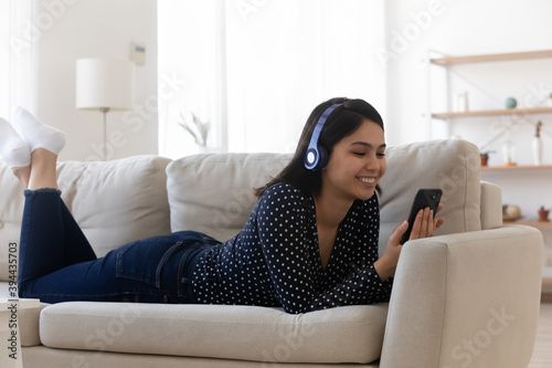 Smiling relaxed Asian young woman wearing headphones using phone, lying on cozy couch in living room, happy satisfied attractive female chatting or shopping online, listening to music, browsing apps
