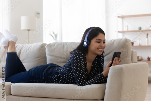 Free Download Smiling relaxed Asian young woman wearing headphones using phone, lying on cozy couch in living room, happy satisfied attractive female chatting or shopping online, listening to music, browsing apps - GFXLoad