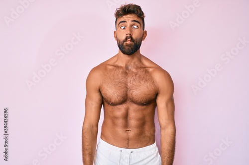 Young hispanic man standing shirtless making fish face with lips  crazy and comical gesture. funny expression.